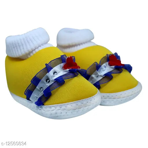 The image shows a pair of infant booties in yellow, which appear to be adorned with a ribbon and possibly some flowers or bows. They are designed for children aged 0 to 12 months