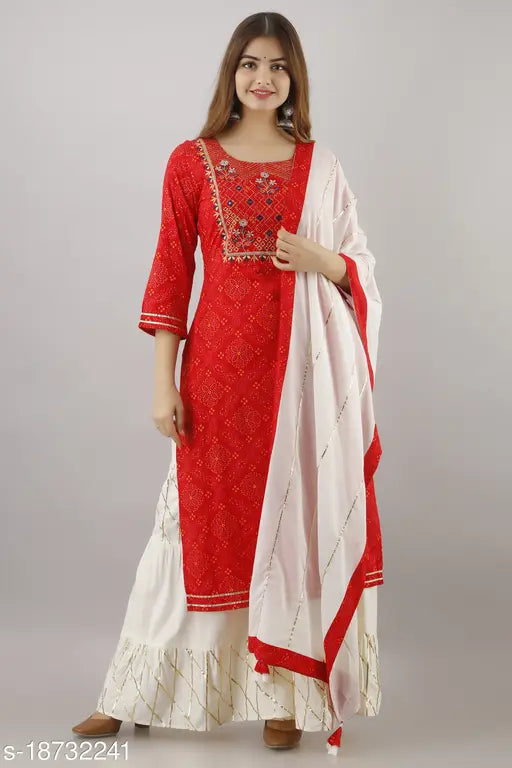 The image is of a woman wearing a vibrant red kurta with long sleeves, a high neckline, and traditional sharara cut. The kurta features intricate rayon embroidery throughout its design, including on the bodice, arms, and dupatta, which appears to be matching in color. The woman is also wearing a long skirt that complements her outfit. The kurta has a straight neckline with what looks like a kameez peeking out from underneath. There are no visible closure or pocket details on the kurta or dupatta