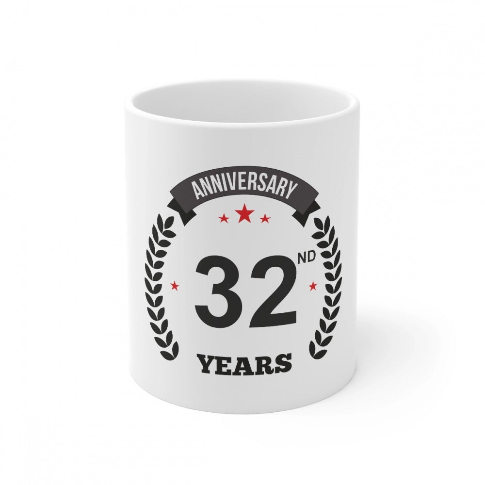 The coffee mug features a graphic and text. It is a white ceramic mug with a 3D printed design. The mug has the phrase "Anniversary 32 years" printed on it, along with a laurel wreath. The visible handle of the mug appears to be made of the same material as the body. The background is not described in the provided image