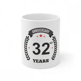 The coffee mug features a graphic and text. It is a white ceramic mug with a 3D printed design. The mug has the phrase "Anniversary 32 years" printed on it, along with a laurel wreath. The visible handle of the mug appears to be made of the same material as the body. The background is not described in the provided image