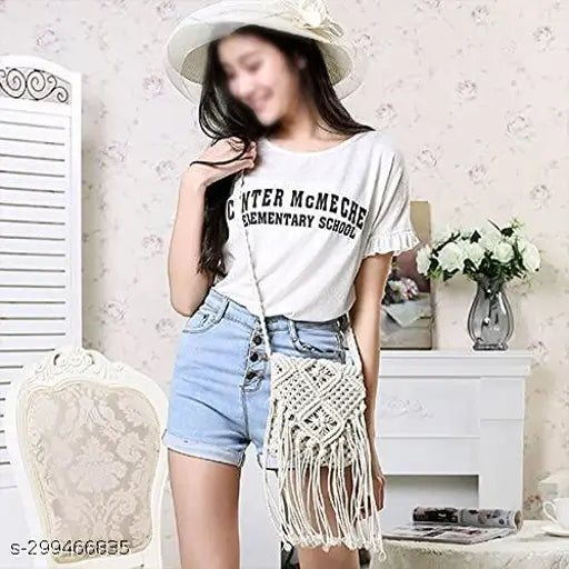 Woman wearing cropped jean shorts and a white t-shirt with text, standing in front of a decorative wall. She's holding a small macramé purse with fringe details
