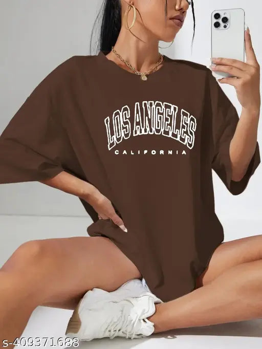 The image features a woman posing with a casual vibe. She is wearing a large, loose-fitting brown sweatshirt with the word "LOS ANGELES" prominently displayed on it. Her pose suggests she might be promoting or showing off the t-shirt