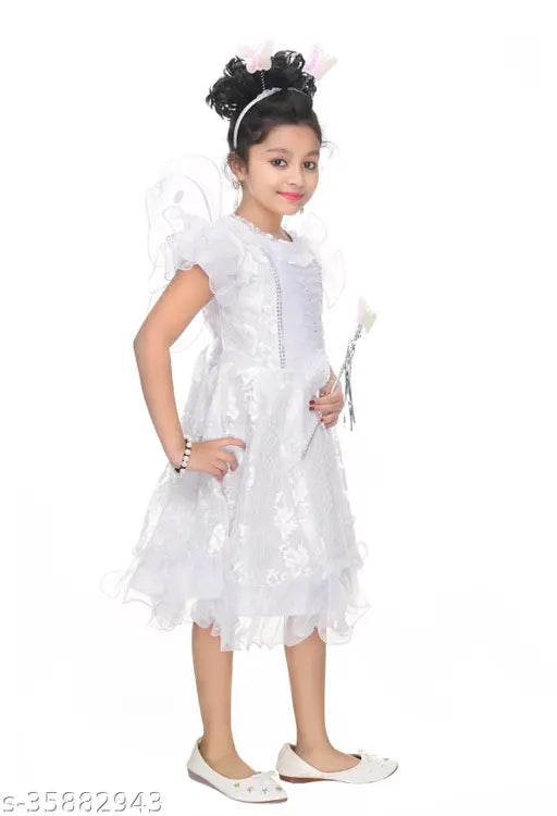 A young girl standing against a white background in a sleeveless white dress with ruffles and a floral pattern, holding a teddy bear