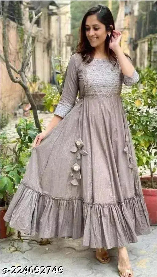 The image displays a woman wearing an off-the-shoulder, long-sleeved grey kameez with puff sleeves and embellished bottom. She stands in front of a building with greenery and a red brick wall