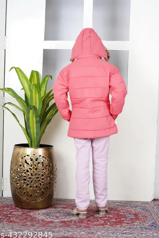 A person in a pink jacket, standing indoors by a window, looking out at a plant