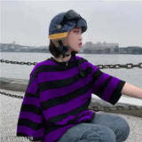 The image features a person in an oversized t-shirt with a fashionable, printed pattern. The shirt appears to have a casual yet trendy style. It's worn by someone who is seated and looking directly at the camera. The person seems to be wearing jeans underneath the t-shirt and has short hair. The background suggests an outdoor setting with a waterfront or dock visible in the distance, where a body of water can be seen. There are no other objects or people clearly visible in the image