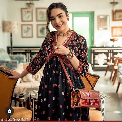 Woman in printed anarkali kurti, black purse, standing indoors with chair and table