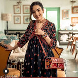 Woman in printed anarkali kurti, black purse, standing indoors with chair and table