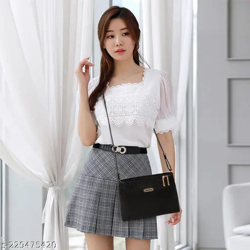 The image shows a woman standing indoors, holding a black shoulder bag with a gold chain strap. She is wearing a white blouse and a dark skirt, accessorized with earrings