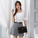 The image shows a woman standing indoors, holding a black shoulder bag with a gold chain strap. She is wearing a white blouse and a dark skirt, accessorized with earrings