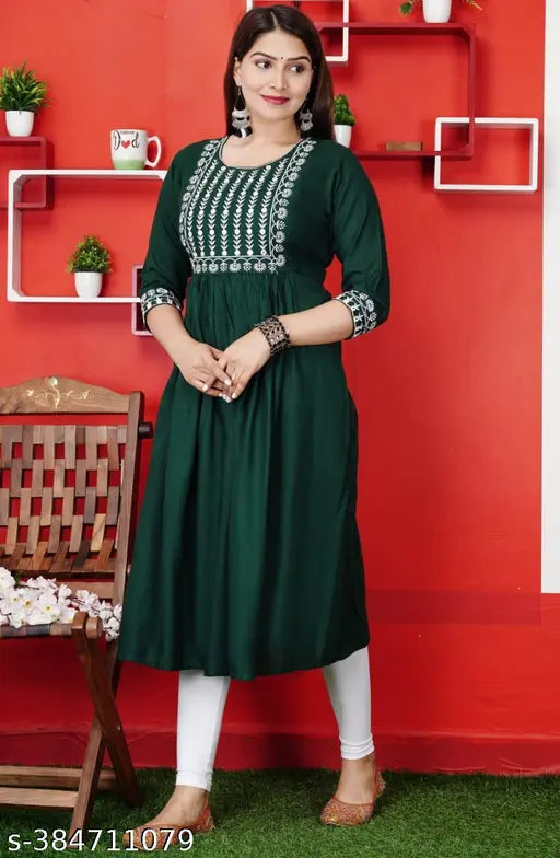The image displays a woman wearing a dark green embroidered kameez with a white dupatta. The outfit has a traditional design, featuring intricate hand-worked embroidery on the bodice and sleeves. The kameez appears to have long sleeves and a full length, with a high neckline. It is paired with white pants or leggings that are not fully visible in the image. In the background, there is a red wall adorned with some decorative items. There is no text visible in the image to provide additional information