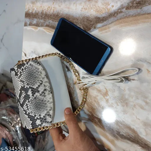 This is an image of a trendy, classy women's slingbag. It features a gold chain strap and a snake skin pattern on the bag. The slingbag appears to be made of a luxurious material with a shiny finish. The bag has a visible closure mechanism and a side pocket that can be used to carry smaller items. The image shows the bag with a smartphone placed inside, demonstrating its utility as a functional accessory