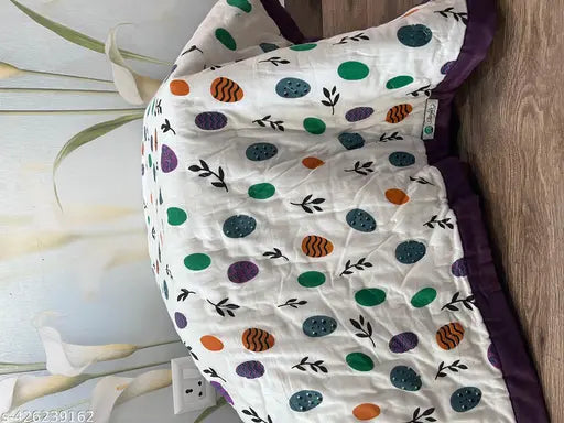 The image features a vibrant, colorful kids quilt with a pattern that includes shapes and spots. It appears to be handmade or crafted, featuring a variety of colors and patterns on what looks like a polyester fabric. The quilt is draped over the side of a piece of furniture, and the visible part has a sizeable enough to suggest it's suitable for a child's bed. The style of the image seems casual and homey, with the focus being on showcasing the quilt itself
