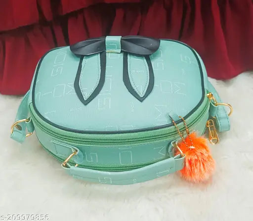 The image shows a fashionable sling bag with a teal or turquoise main body and a light blue or sky blue accented section. It features a bow-like closure detail and an adjustable strap. A small tassel is attached to one of the straps, contributing to the bag's decorative details. The brand label appears to be visible on the lower front part of the bag