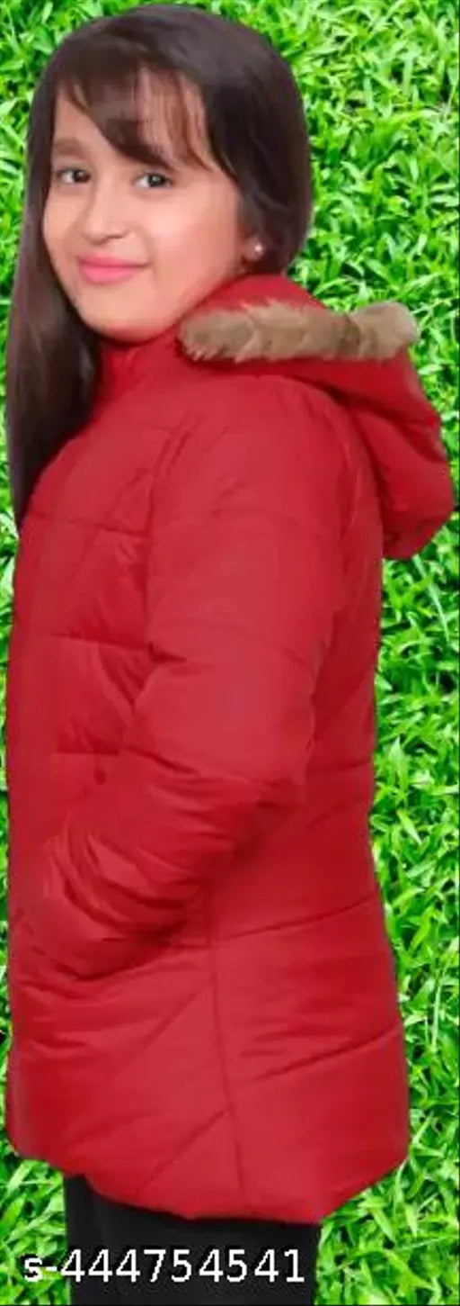 This is an image of a young girl wearing a red padded jacket, standing outdoors. The jacket features a hood and has a fur-lined collar for warmth. The background is blurred with a green hedge, giving a sense of a garden or park setting