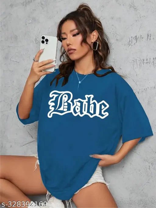 A woman in a blue and white baggy t-shirt with a printed logo standing against a grey brick wall, holding a phone