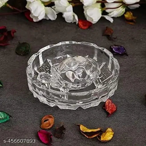 The crystal plate is empty and features a turtle motif on top. It sits against a gray surface, surrounded by small, colorful leaves or petals