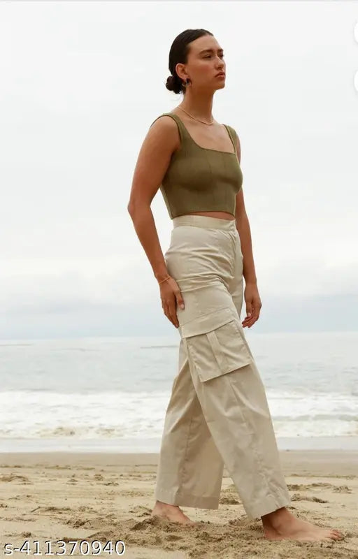 The image is of a woman wearing a green crop top and cream cargo pants. She appears to be on a beach, standing near the water's edge. The pants have four pockets visible and are styled in an oversized, baggy fashion typical of cargo pants