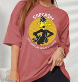 The oversized round neck t-shirt features a vibrant graphic design on a maroon background, prominently displaying a cartoon character with text that reads "Sarcasm Just Another Service Offer". This fashionable piece of clothing is perfect for women and girls who appreciate humor