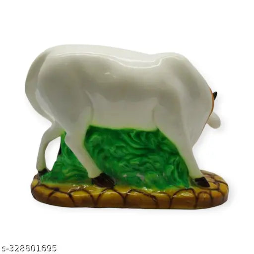 This is an image of a white marble dust cow statue. The cow statue is designed to appear as if it has green grass under its feet, with the marble dust creating this effect. The cow is depicted in a standing position and features simple, unadorned details that give it a minimalist and decorative appearance. It appears to be a small ornamental item suitable for home or office decoration and potentially as a gift item