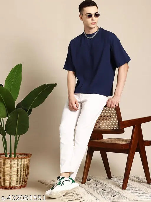 A man wearing a white jean with rolled-up cuffs, a navy blue shirt, and sunglasses. He's posing in front of a potted plant against a light-colored wall